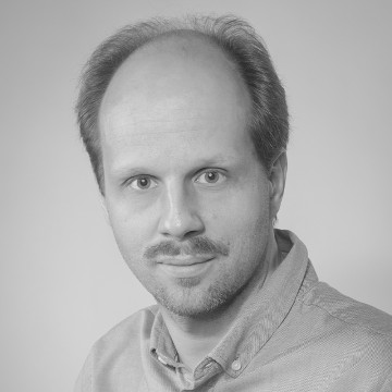 Miehellä, jolla on taantuva hiusraja ja lyhyet kasvohiukset, on vaalea napillinen paita, ja hän katsoo neutraalilla ilmeellä suoraan kameraan tavallista taustaa vasten. Kuva on mustavalkoinen.