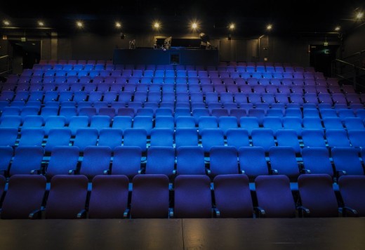 Sinisten ja violettien teatteripenkkien tyhjät rivit näkyvät hämärässä salissa, joka on näyttämölle päin, ja valonheittimet valaisevat istuinalueen ja takana näkyvän ohjauskopin.