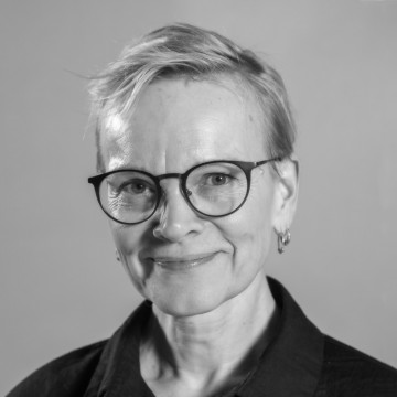 Mustavalkoinen muotokuva henkilöstä, jolla on lyhyet hiukset, pyöreät silmälasit, pienet korvakorut ja kauluspaita, hymyilee hieman ja katsoo suoraan kameraan tavallista taustaa vasten.