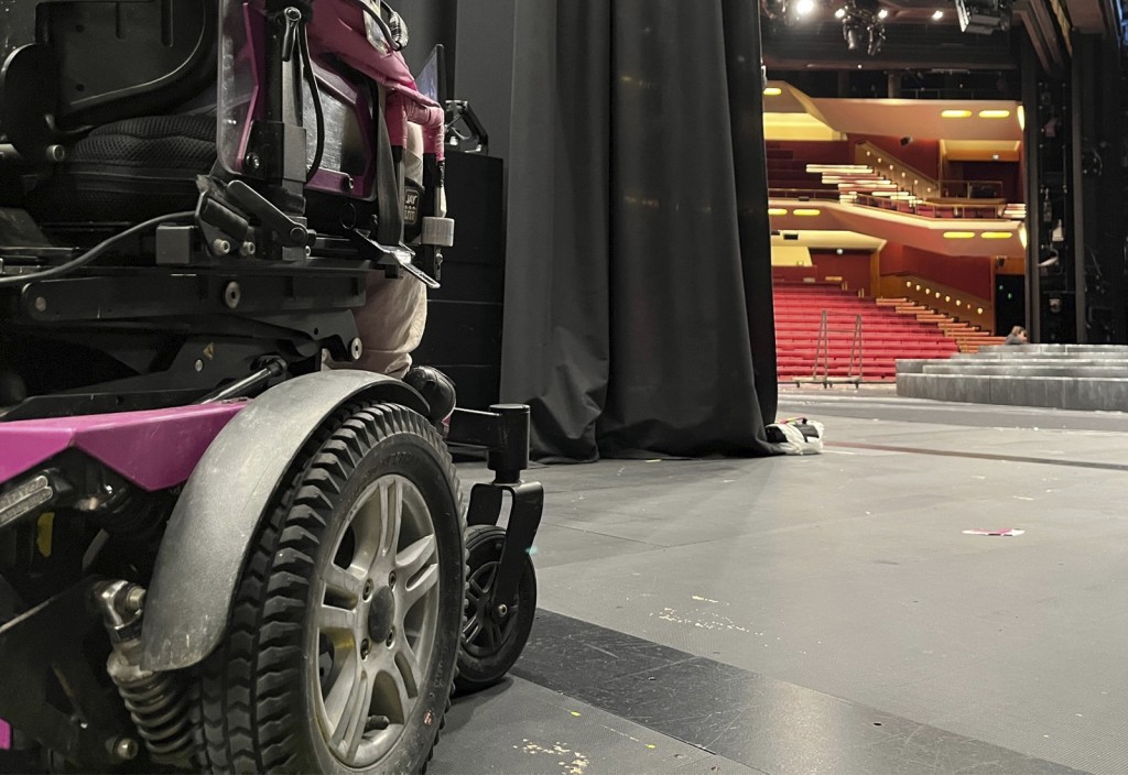 Lähikuva vaaleanpunaisesta sähköpyörätuolista teatterin lavalla, taustalla näkyy verhot ja punaiset tyhjät katsomon istuimet.
