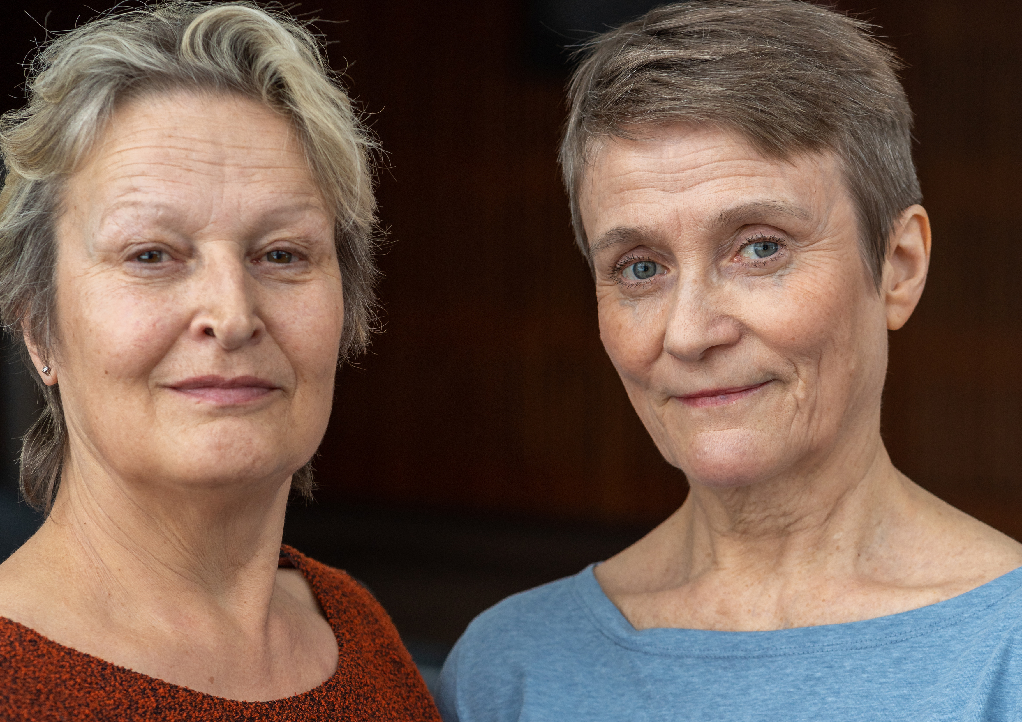 Kaksi vanhempaa, lyhythiuksista naista seisoo lähekkäin ja katsoo kameraan lempein ilmein. Toisella on yllään oranssi pusero, toisella vaaleansininen toppi tummaa, epäselvää taustaa vasten.