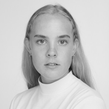 Mustavalkoinen muotokuva nuoresta henkilöstä, jolla on pitkät, suorat hiukset, päällään vaalea poolokaulus ja pienet korvakorut, katselee suoraan kameraan neutraali ilme tavallista taustaa vasten.