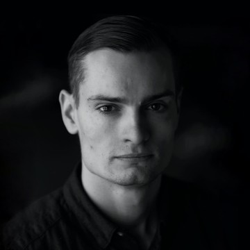 Mustavalkoinen muotokuva nuoresta miehestä, jolla on lyhyet, siististi kammatut hiukset, yllään tumma kauluspaita, katselee suoraan kameraan neutraali ilme tummaa taustaa vasten.