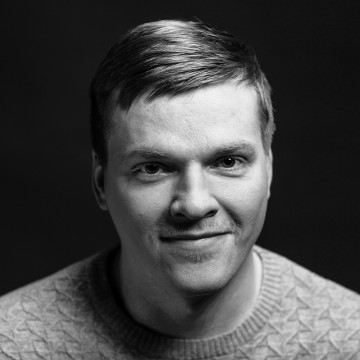 Lyhyet vaaleat hiukset omaava mies, jolla on yllään kuvioitu villapaita, katsoo suoraan kameraan ja hymyilee hieman. Muotokuva on mustavalkoinen ja siinä on tumma tausta.