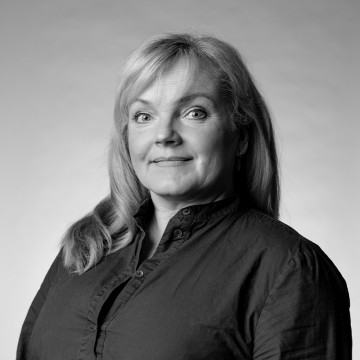Mustavalkoinen muotokuva vaaleahiuksisesta naisesta, jolla on napillinen paita, kasvot hieman sivulle päin ja hymyilee pehmeästi. Tausta on tavallinen ja pehmeästi valaistu.