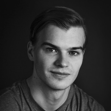 Nuori mies, jolla on lyhyet, siististi kammatut hiukset ja hento hymy, katsoo suoraan kameraan. Hänellä on yllään yksinkertainen T-paita, ja tausta on tumma, mikä antaa muotokuvalle klassisen, tunnelmallisen tunnelman.