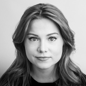 Nuori nainen, jolla on aaltoilevat, olkapään pituiset hiukset, katsoo suoraan kameraan neutraalilla ilmeellä. Kuva on mustavalkoinen, ja nainen on sijoitettu tavallista taustaa vasten.
