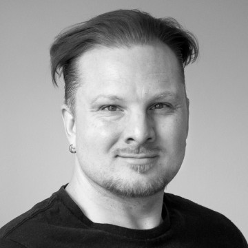 Vaalea mies, jolla on vaalea iho, lyhyet liukuhiukset, lyhyt parta ja viikset, hymyilee kevyesti, kun hänellä on musta paita ja pieni korvarengas, seisoo vaalean taustan edessä.