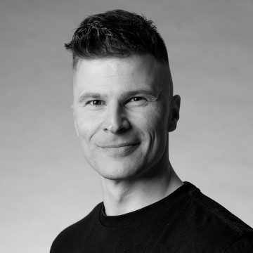 Mustavalkoinen muotokuva henkilöstä, jolla on lyhyet, muotoillut hiukset, yllään tavallinen tumma t-paita, hymyilee hieman ja katsoo suoraan kameraan tavallista taustaa vasten.