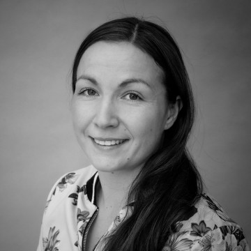 Mustavalkoinen muotokuva naisesta, jolla on pitkät tummat hiukset ja joka hymyilee kameralle. Hänellä on yllään kukkakuvioinen toppi ja hän poseeraa tavallista taustaa vasten.