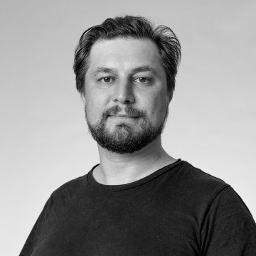 Lyhyet tummat hiukset ja partainen mies, jolla on musta t-paita, poseeraa vaaleaa taustaa vasten. Kuva on mustavalkoinen, ja mies katsoo rauhallisesti kameraan.