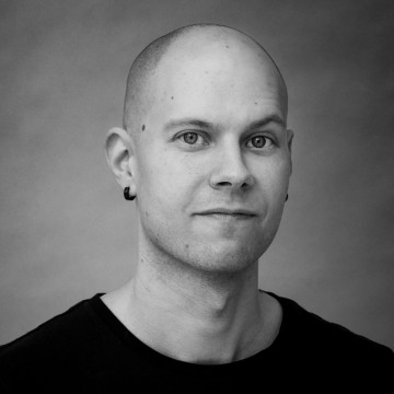 Vaaleaihoinen kalju mies, jolla on musta paita ja pienet korvakorut, katsoo kameraan neutraalilla ilmeellä. Tausta on tavallinen ja pehmeästi valaistu. Kuva on mustavalkoinen.