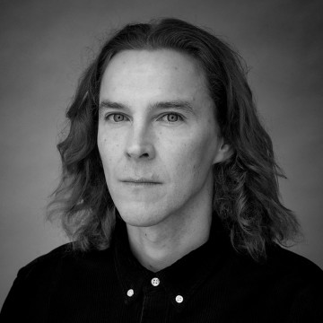 Mustavalkoinen muotokuva henkilöstä, jolla on hartioiden pituinen aaltoileva tukka, päällään napitettu tumma paita, katselee suoraan kameraan neutraali ilme tavallista taustaa vasten.