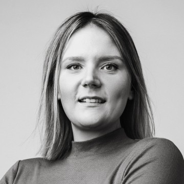 Mustavalkoinen muotokuva nuoresta naisesta, jolla on suorat, vaaleat hiukset ja pitkähihainen, korkeakaulainen yläosa. Hän katsoo kameraan ja hymyilee kevyesti, taustana tavallinen tausta.