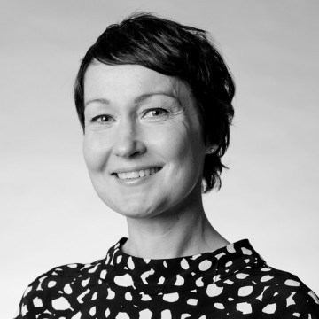 Mustavalkoinen muotokuva hymyilevästä naisesta, jolla on lyhyet tummat hiukset, päällään kuviollinen toppi, kameraan päin, neutraali tausta.