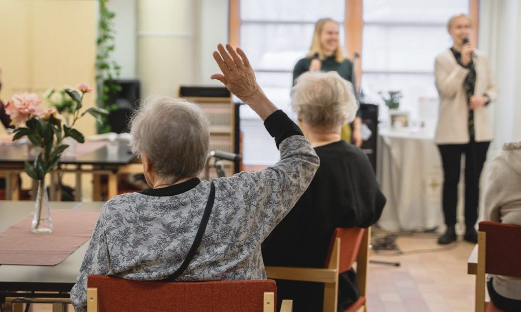 Iäkäs henkilö nostaa kätensä, kun hän istuu muiden joukossa yhteishuoneessa ja kuuntelee kahden naisen puhetta tai laulua edessä. Pöydillä on maljakoita, joissa on kukkia.