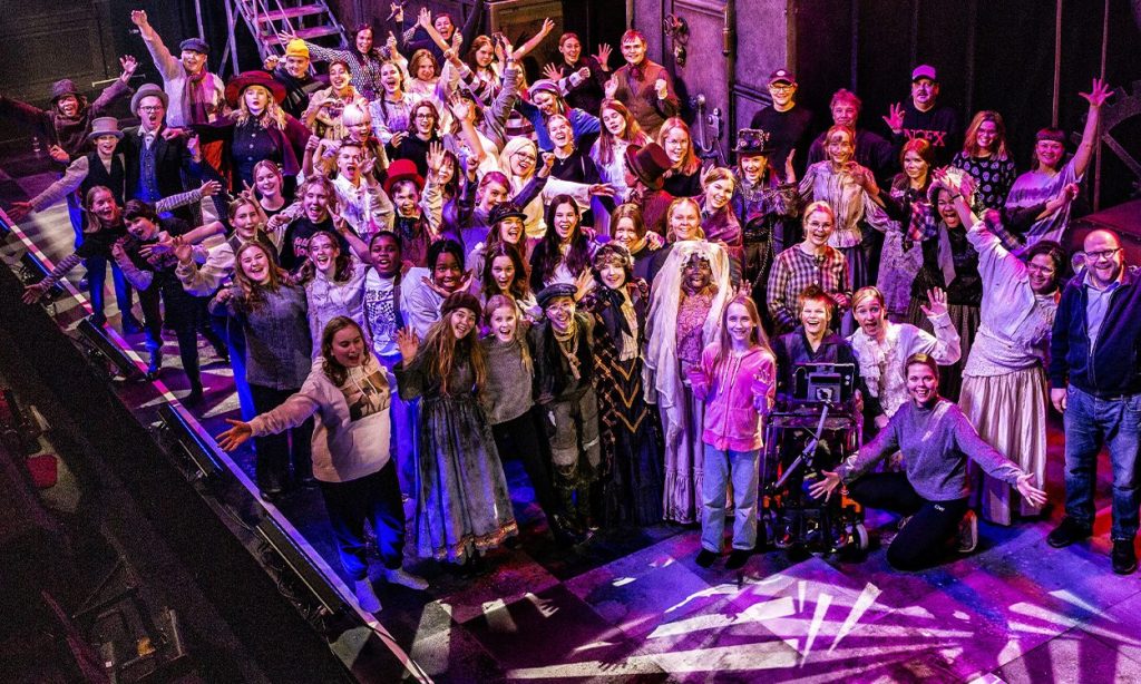 Suuri joukko värikkäisiin asuihin pukeutuneita ihmisiä poseeraa yhdessä teatterin lavalla hymyillen ja kädet kohotettuna, näyttämötarvikkeiden ja dramaattisen valaistuksen ympäröimänä.