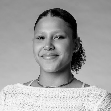 Mustavalkoinen muotokuva nuoresta henkilöstä, jolla on taaksepäin kiharat hiukset, nenärengas, korvakorut, ketjukoru ja kevyt neuletoppi, ja joka hymyilee pehmeästi tavallista taustaa vasten.