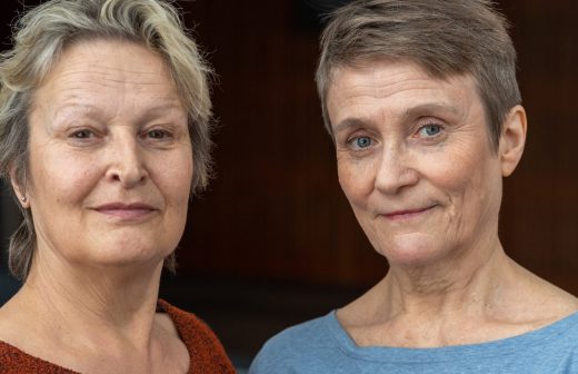 Kaksi vanhempaa naista, joilla on lyhyet harmaat hiukset, seisovat lähekkäin ja katsovat kameraan lempein ilmein. Toisella on yllään oranssi villapaita ja toisella vaaleansininen toppi, molemmat pehmeästi valaistussa sisätilassa.