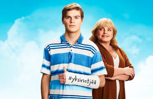 Nuori mies siniraidallisessa poolopaidassa, jonka kädessä on kipsattu teksti #yksinetjää, seisoo hymyilevän ruskeaan neuletakkiin pukeutuneen naisen vieressä, molemmat kirkkaan sinistä taivasta vasten.