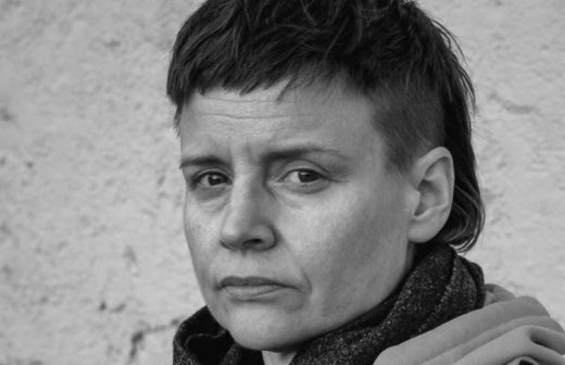 Mustavalkoinen muotokuva henkilöstä, jolla on lyhyet tummat hiukset ja vakava ilme ja joka katsoo hieman kameran ohi. Päällään huivi ja takki, taustalla kuvioitu seinä.