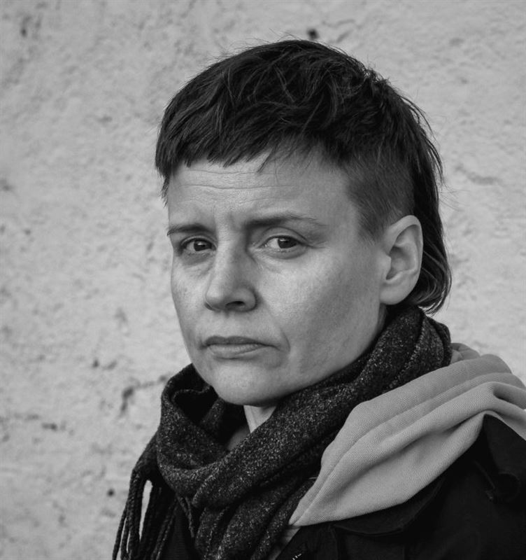 Mustavalkoinen valokuva henkilöstä, jolla on lyhyet, kuvioidut hiukset ja vakava ilme, päällään huivi ja takki, ja joka seisoo karhean, vaalean seinän edessä.