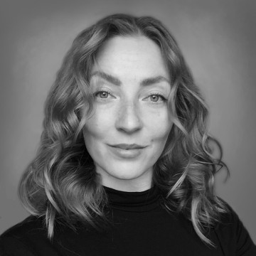 Henkilö, jolla on aaltoilevat, olkapään pituiset hiukset ja vaaleat silmät, katsoo kameraan hymyillen, ja hänellä on yllään musta poolopusero harmaata taustaa vasten. Kuva on mustavalkoinen.