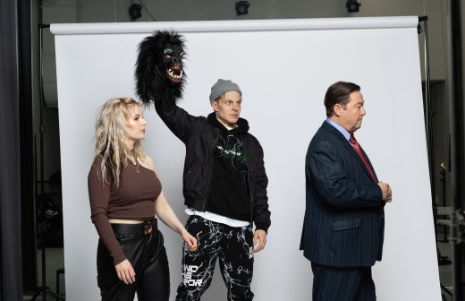 Kolme ihmistä seisoo valkoisen taustan edessä. Ruskeaan toppiin pukeutunut nainen katsoo vasemmalle, rennosti pukeutunut mies pitää mustaa gorillanaamiota päänsä yläpuolella, ja pukuun pukeutunut mies katsoo oikealle.