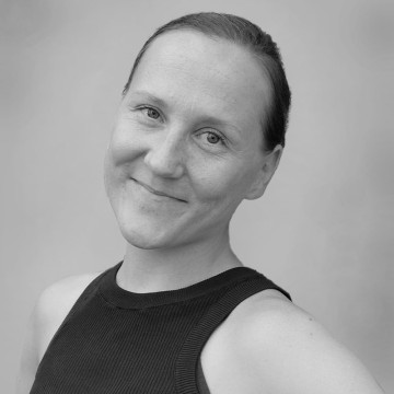 Henkilö, jolla on vaalea iho ja vaaleat silmät, hymyilee hellästi, hänellä on hihaton musta toppi ja hän seisoo vaaleanharmaalla taustalla. Kuva on mustavalkoinen.