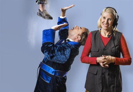 Siniseen samettitakkiin pukeutunut mies näyttelee dramaattisesti, kun hänen yläpuolellaan roikkuu pari jalkaa tossuissa. Hänen vieressään seisoo hymyilevä vaaleahiuksinen nainen, jolla on kuulokkeet ja harmaa liivi, kädet ristissä. Sininen tausta.