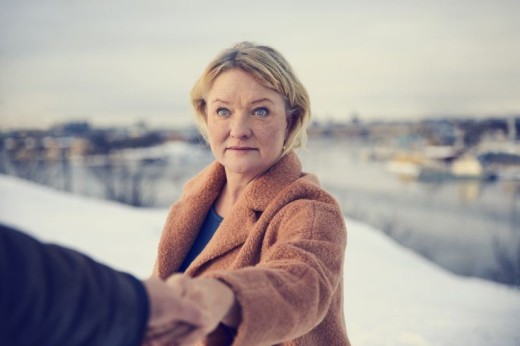 Ruskeaan takkiin pukeutunut nainen seisoo ulkona lumisessa maisemassa veden äärellä, katsoo eteenpäin ja ojentaa samalla kätensä jonkun kädestä. Taustalla näkyy veneitä ja rakennuksia.