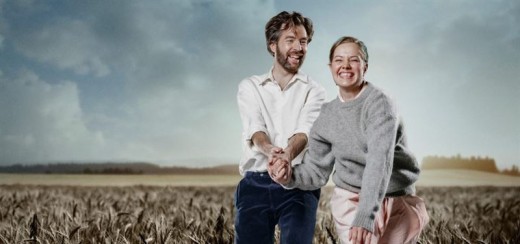 Hymyilevä mies ja nainen kädestä pitäen ja nauraen seisovat korkealla viljapellolla pilvisen taivaan alla. He ovat pukeutuneet rennosti ja vaikuttavat onnellisilta, ja taustalla näkyy avoin maaseutu.