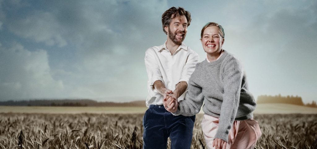Mies ja nainen seisovat vehnäpellolla kädestä pitäen ja hymyillen iloisesti kameralle. Taivas on pilvinen, ja molemmilla on rennot vaatteet, ja he vaikuttavat iloisilta ja rennoilta ulkona.