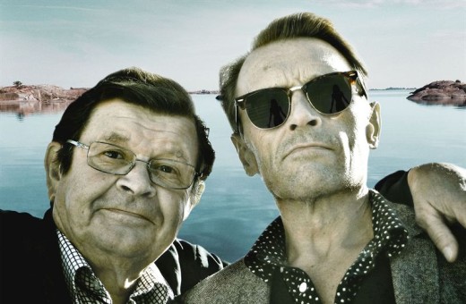 Kaksi miestä poseeraa selfietä ottaessaan rauhallisella järvellä, jonka rannat ovat kivikkoisia. Toisella miehellä on silmälasit ja hän hymyilee, kun taas toisella on aurinkolasit ja hänellä on käsi ensimmäisen miehen ympärillä. Taivas on kirkas.