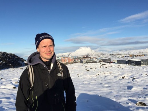 Mustaan takkiin ja pipoon pukeutunut mies seisoo lumisessa maisemassa, jonka taustalla on kaupunki ja lumipeitteinen vuori kirkkaan sinisen taivaan alla.