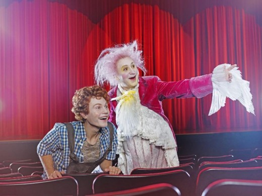 Kaksi värikkäisiin teatteripukuihin pukeutunutta ihmistä istuu teatterin istuimissa. Toinen, jolla on kiharat hiukset ja ruutupaita, hymyilee innoissaan. Toinen, jolla on vaaleanpunainen peruukki ja hienostunut mekko, osoittaa suurieleisesti kohti näyttämöä, jonka taustalla on punaiset verhot.