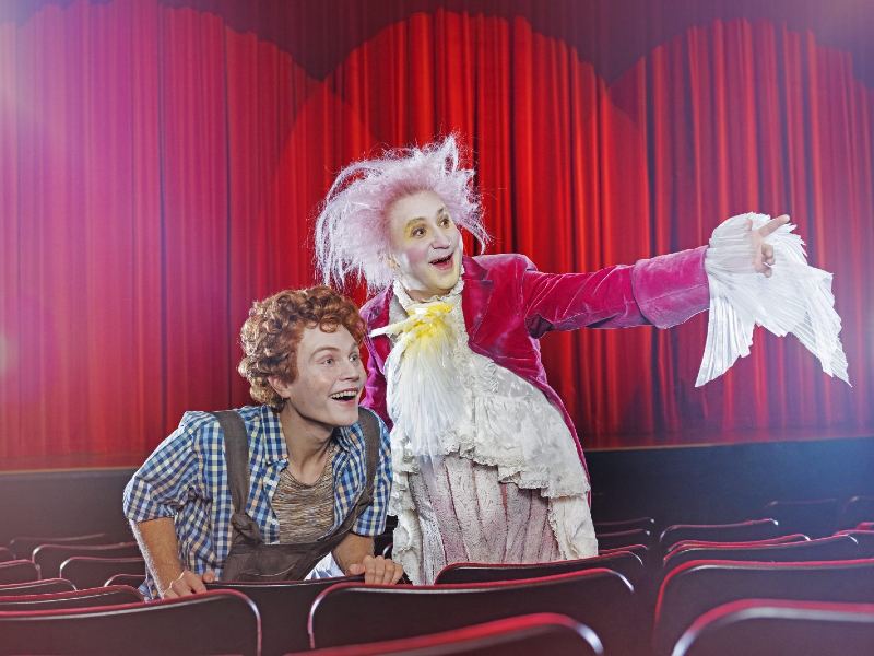 Kaksi värikkäisiin teatteripukuihin pukeutunutta näyttelijää istuu teatterissa. Toisella on kiharat punaiset hiukset ja ruudullinen paita, kun taas toisella on kirkkaan vaaleanpunainen takki ja valkoinen peruukki, ja hän hymyilee ja osoittaa dramaattisesti kohti näyttämöä.