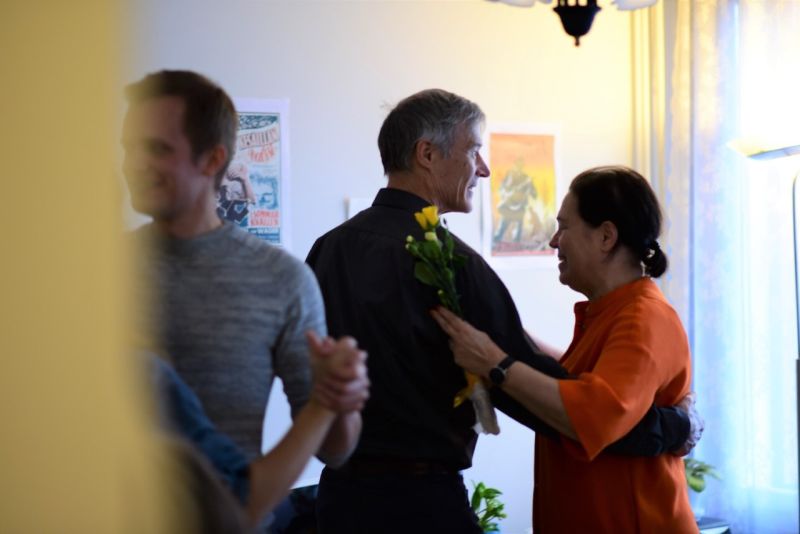 Vanhempi pariskunta tanssii yhdessä sisätiloissa hymyillen, kun oranssiin toppiin pukeutunut nainen pitelee keltaisia kukkia. Vasemmalla näkyy toinen tanssiva pariskunta, ja taide-esineet koristavat seinää taustalla.