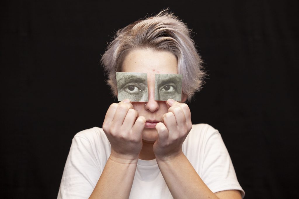 Henkilö, jolla on lyhyet vaaleat hiukset, pitää omia silmiään painettujen silmien pieninä paperileikkeinä, mikä luo illuusion erilaisista silmistä. Hänellä on yllään valkoinen paita ja hän poseeraa mustaa taustaa vasten.