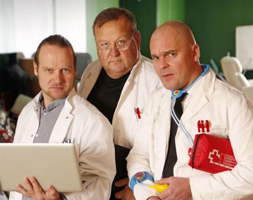 Kolme valkoisiin laboratoriotakkeihin pukeutunutta miestä poseeraa yhdessä sisätiloissa. Yhdellä on kannettava tietokone, toinen seisoo keskellä silmälasit silmillään, ja kolmannella on lelustetoskooppi ja punainen lääkintäpakkaus. He vaikuttavat vakavilta ja keskittyneiltä.