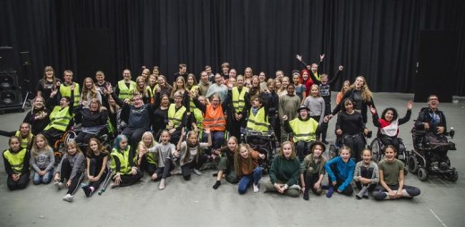 Suuri, erilainen joukko ihmisiä, mukaan lukien pyörätuolissa istuvia ja heijastinliiveihin pukeutuneita henkilöitä, poseeraa yhdessä sisätiloissa hymyillen ja juhlien mustan verhon edessä.