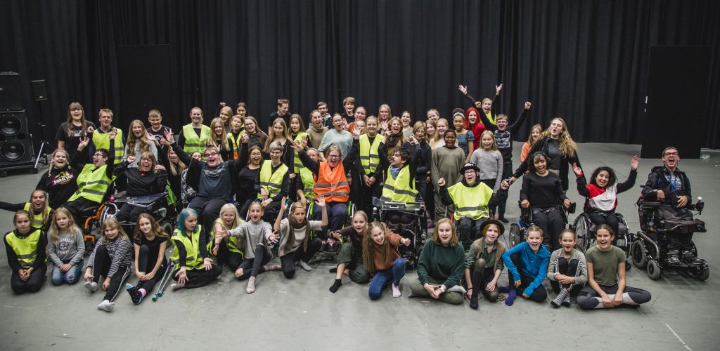 Suuri joukko ihmisiä, mukaan lukien lapsia, aikuisia ja pyörätuolissa istuvia henkilöitä, poseeraa yhdessä ja hymyilee tilavassa sisätilassa. Joillakin on keltaiset turvaliivit, ja taustalla roikkuu musta verho.
