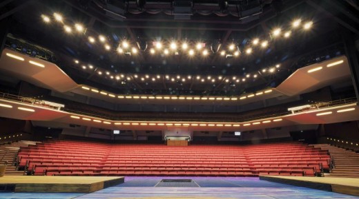 Suuri, moderni teatterisali, jossa on kolme kerrosta tyhjiä punaisia istuimia näyttämöltä katsottuna. Kirkkaat kattovalaisimet valaisevat tilaa, ja seinissä on geometrisia linjoja ja parvekkeita.
