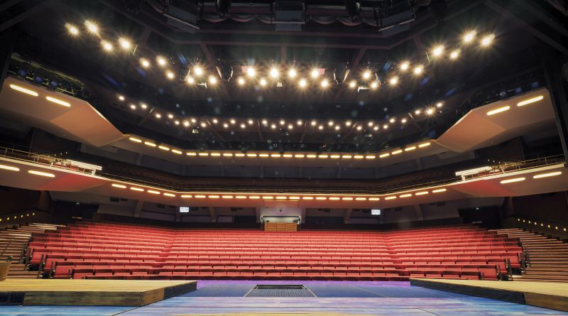 Laaja näkymä teatterin näyttämöltä, jossa on riviin sijoitetut tyhjät punaiset istuimet, joiden yläpuolella on kaksi parveketta ja lukuisat valot, jotka valaisevat salin.