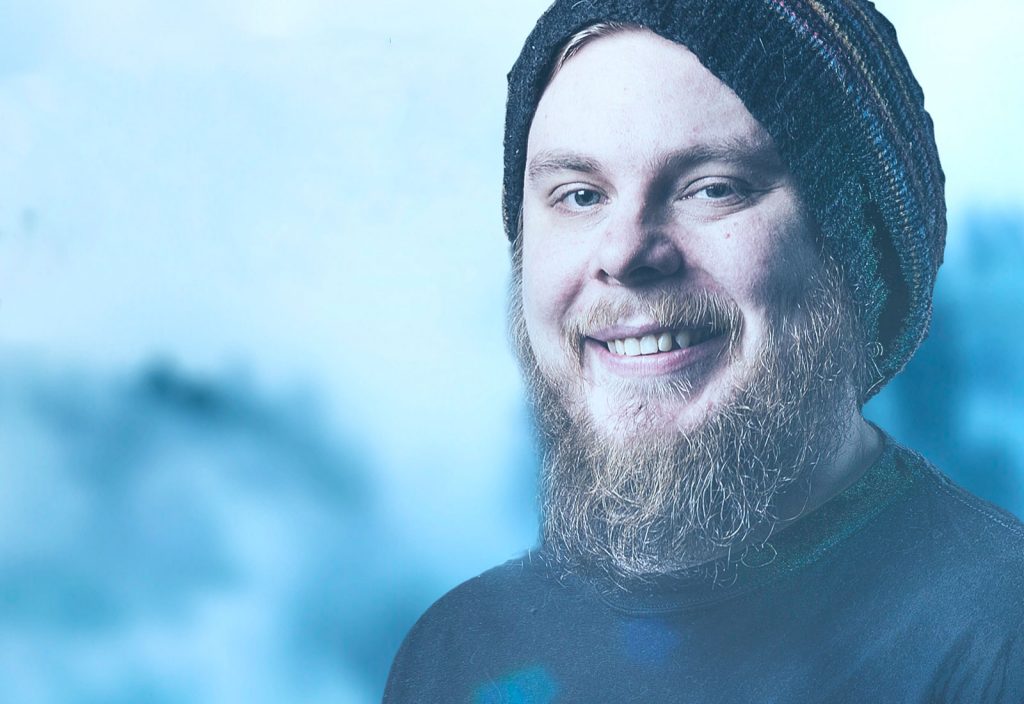 Hymyilevä nuori mies, jolla on täysparta ja viikset, päällään raidallinen neuletakki ja tumma paita, seisoo sinistä, sumeaa taustaa vasten.