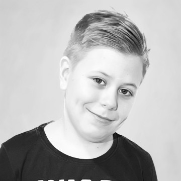 Nuori poika, jolla on vaaleat, sivulle tyylitellyt hiukset, hymyilee hellästi, yllään tumma paita ja poseeraa mustavalkoisessa valokuvassa tavallista, vaaleaa taustaa vasten.