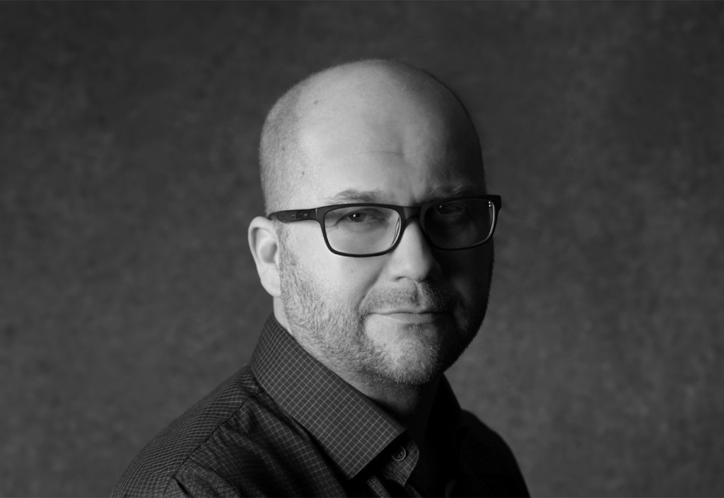 Mies, jolla on kalju pää, silmälasit ja lyhyt parta ja jolla on ruudullinen paita, katsoo mustavalkoisessa muotokuvassa suoraan kameraan tavallista, tummaa taustaa vasten.
