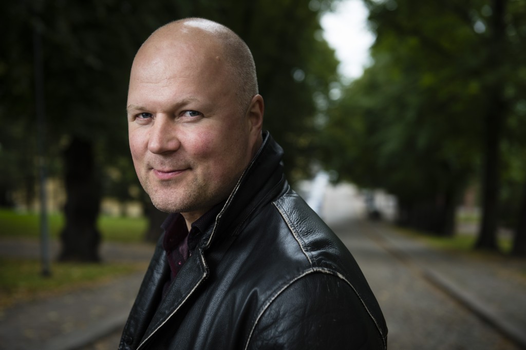 Mustaan nahkatakkiin pukeutunut kalju mies seisoo ulkona puiden reunustamalla polulla ja hymyilee hieman kameralle, jonka vihreä tausta on sumea.