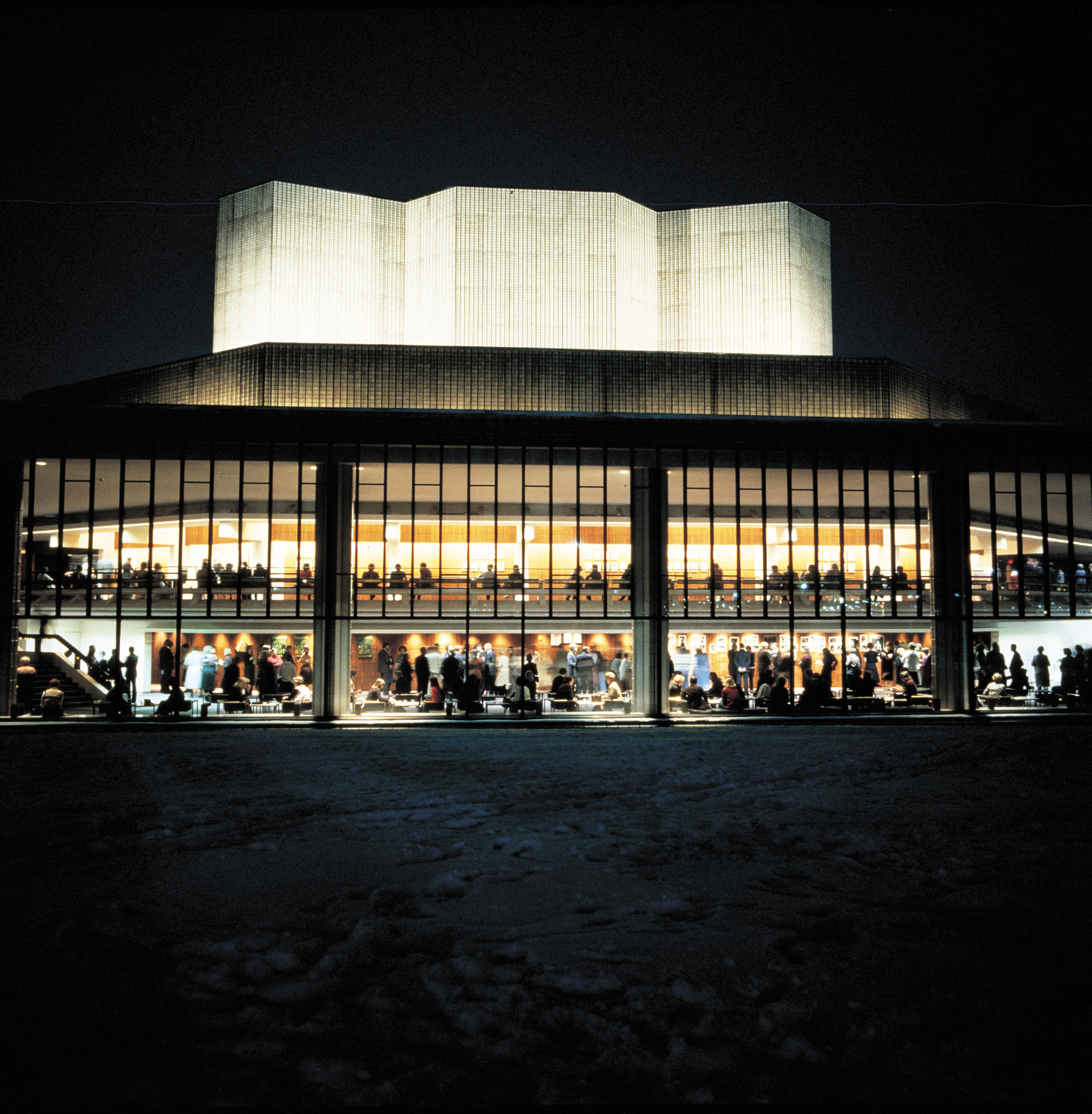 Kirkkaasti valaistu moderni teatterirakennus yöllä, jossa suuret lasi-ikkunat paljastavat kaksi kerrosta täynnä ihmisiä; ulkona on lunta.