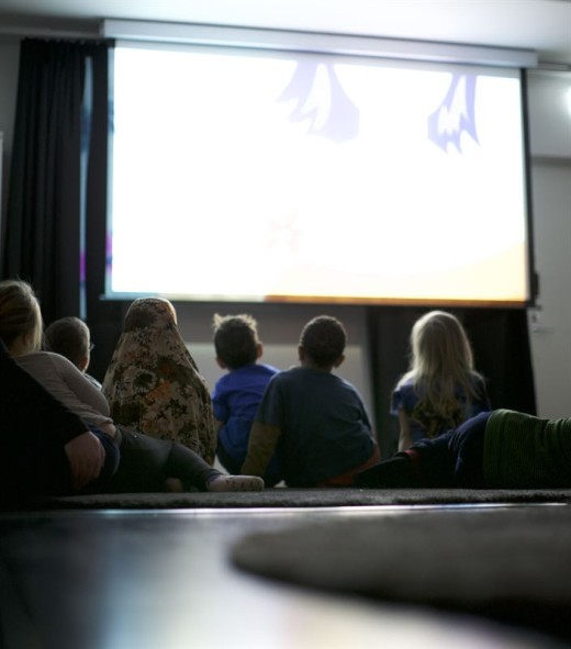 Ryhmä lapsia istuu lattialla, vastapäätä suurta, kirkasta näyttöä hämärässä huoneessa, ja katsoo yhdessä heijastettua elokuvaa tai animaatiota.